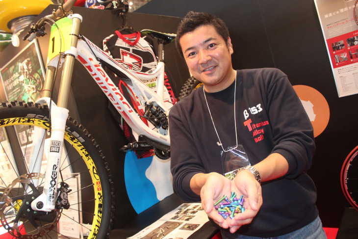 井手川選手のDevinciをバックに社長の柿沢宏一さんが手に持つのはチタンボルトは鮮やかなカラーが特徴の1つ