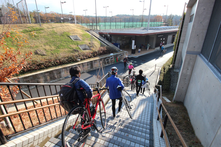 自転車を担いでサイクリングコースへ。奥に見えるのがレンタルサイクルの受付所だ